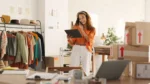 Small business owner using a tablet to manage an online store with clothing inventory and shipping boxes in a workspace